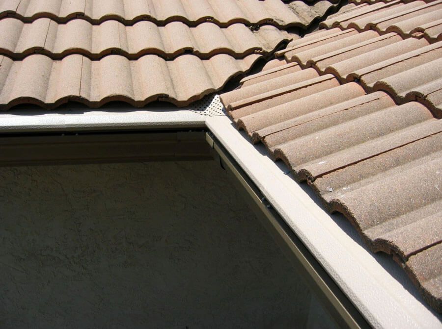 A close-up of a house roof showing tan clay tiles meeting at a corner gutter with a metal edge trim.