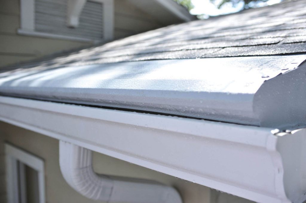 A white metal gutter and downspout attached to the eaves of a house with light gray shingles.