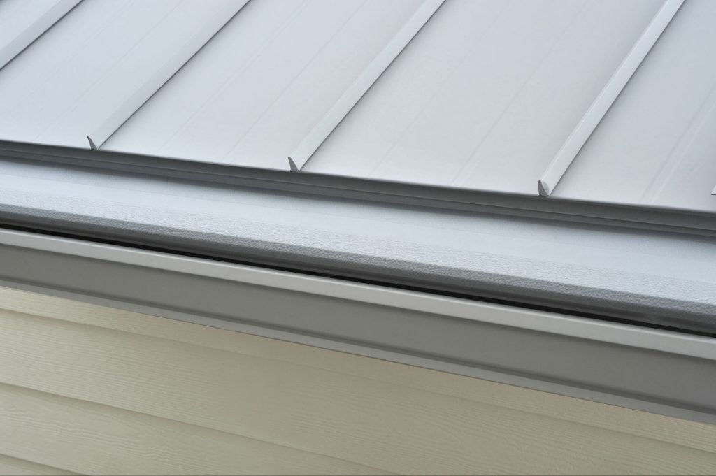 A silver metal roof with standing seams overhangs a gray gutter attached to light tan horizontal siding.
