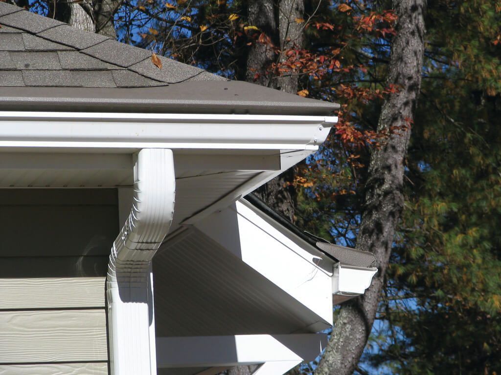 A close-up view of the white metal gutter and downspout system installed on the roof eaves of a house with tan siding.