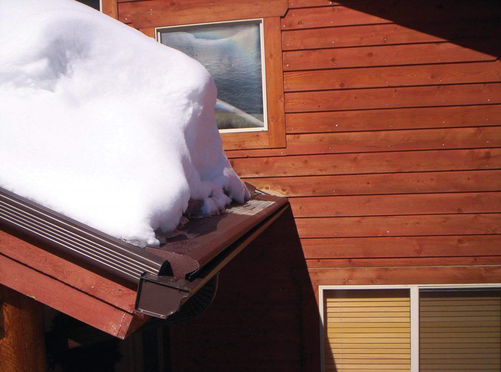 Thick snow covers the roof of a brown log cabin, hanging over the gutter with a window visible on the siding.