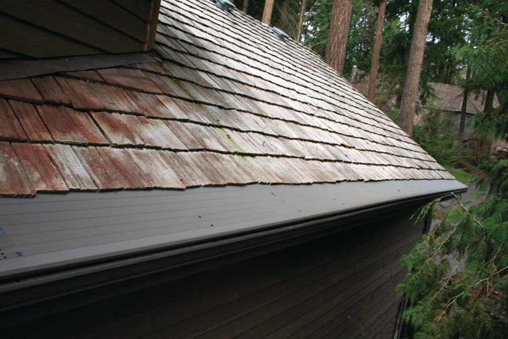 A weathered wood shake roof with signs of moss growth, above a dark horizontal gutter and wooden siding in a forest setting.