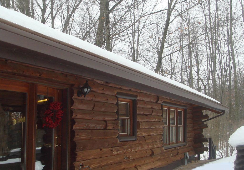 A rustic log cabin exterior during a snowy winter day, featuring dark wood walls, a dark trim, and a festive red wreath.