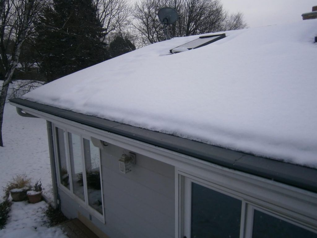 A snow-covered roof with a skylight above a house exterior featuring windows and a wall-mounted light fixture.