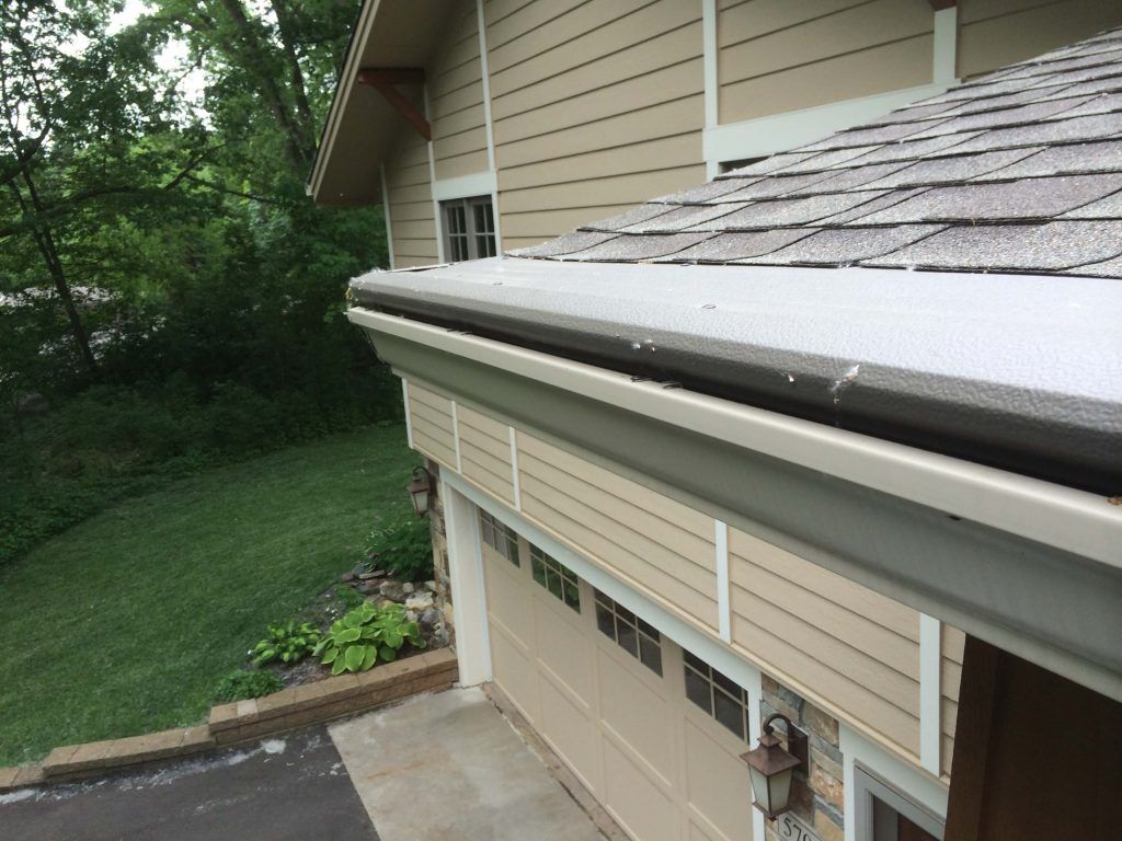 An elevated side view of a house garage featuring tan siding, a shingled roof, and a white gutter with a leaf guard.