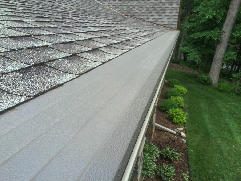 A close-up view of a grey gutter cover installed on a residential roofline, with a lawn and trees visible in the background.