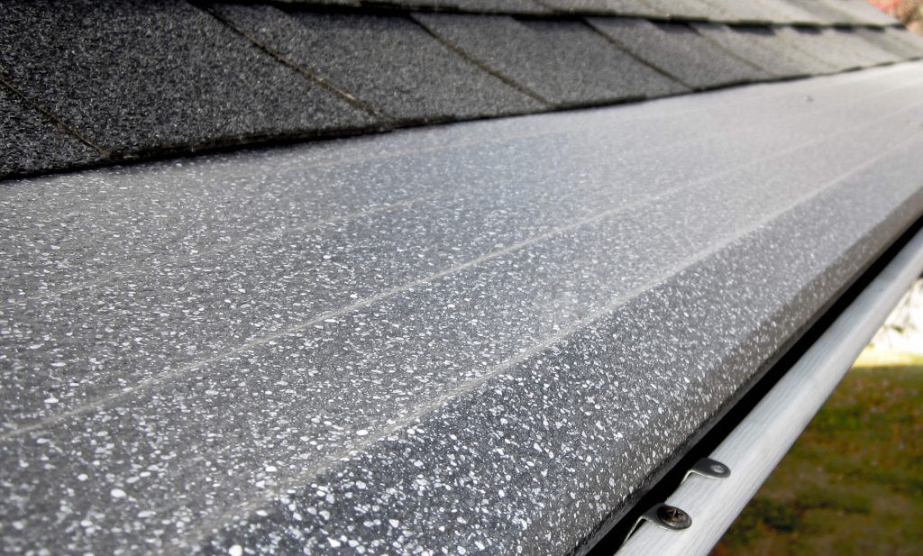 A gray, textured gutter guard installed over a white rain gutter on a roof with dark asphalt shingles.
