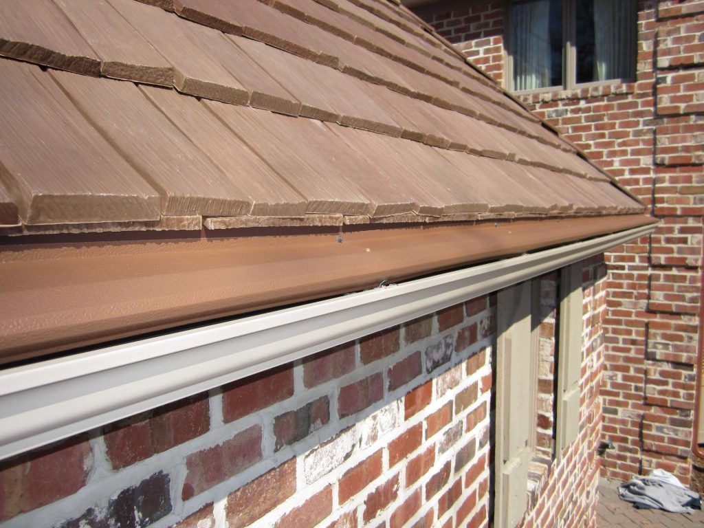 A close-up view of a brown tile roof with a matching gutter above a brick wall and window with beige shutters.