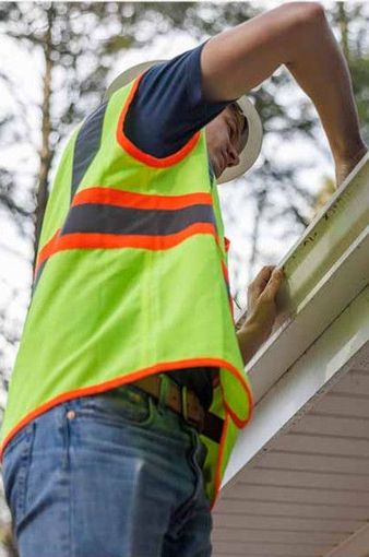 A person in a high-visibility vest and hard hat works on a residential roof gutter.