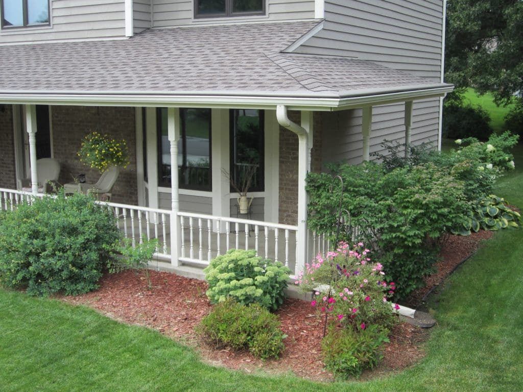 A house with a wraparound porch featuring white railings, a tan siding exterior, and landscaped garden beds with mulch.