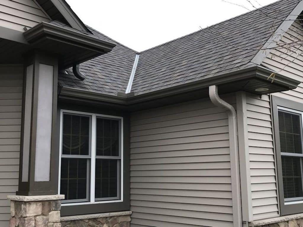 The exterior corner of a house featuring gray vinyl siding, a dark shingled roof, brown trim, and a stone-based pillar.
