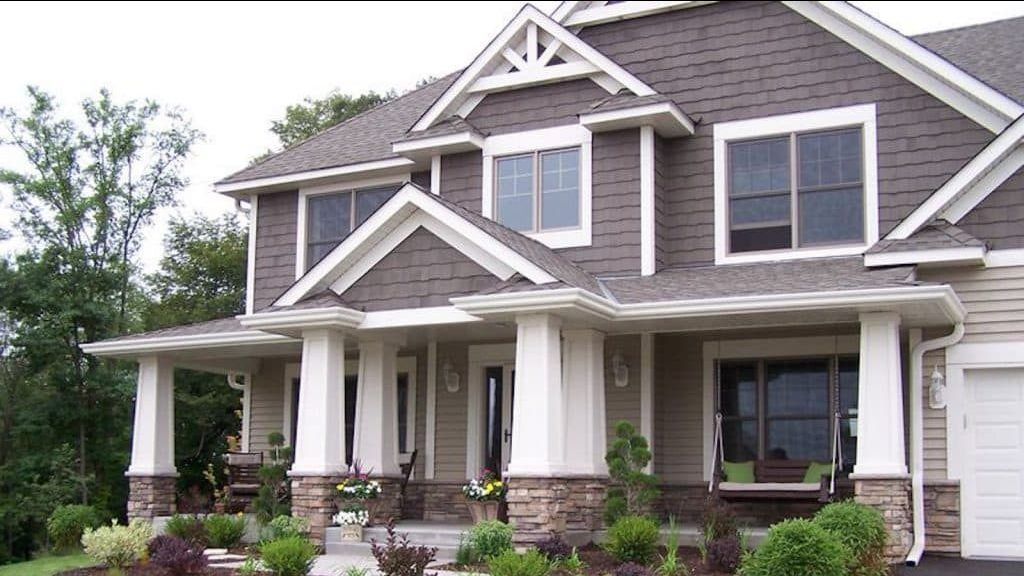 A two-story suburban home with tan siding, gray shingles, white trim, stone accents, and a front porch with white columns.