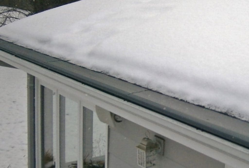 A thick blanket of snow rests on a gray roof edge above a house window with a small light fixture on the wall.
