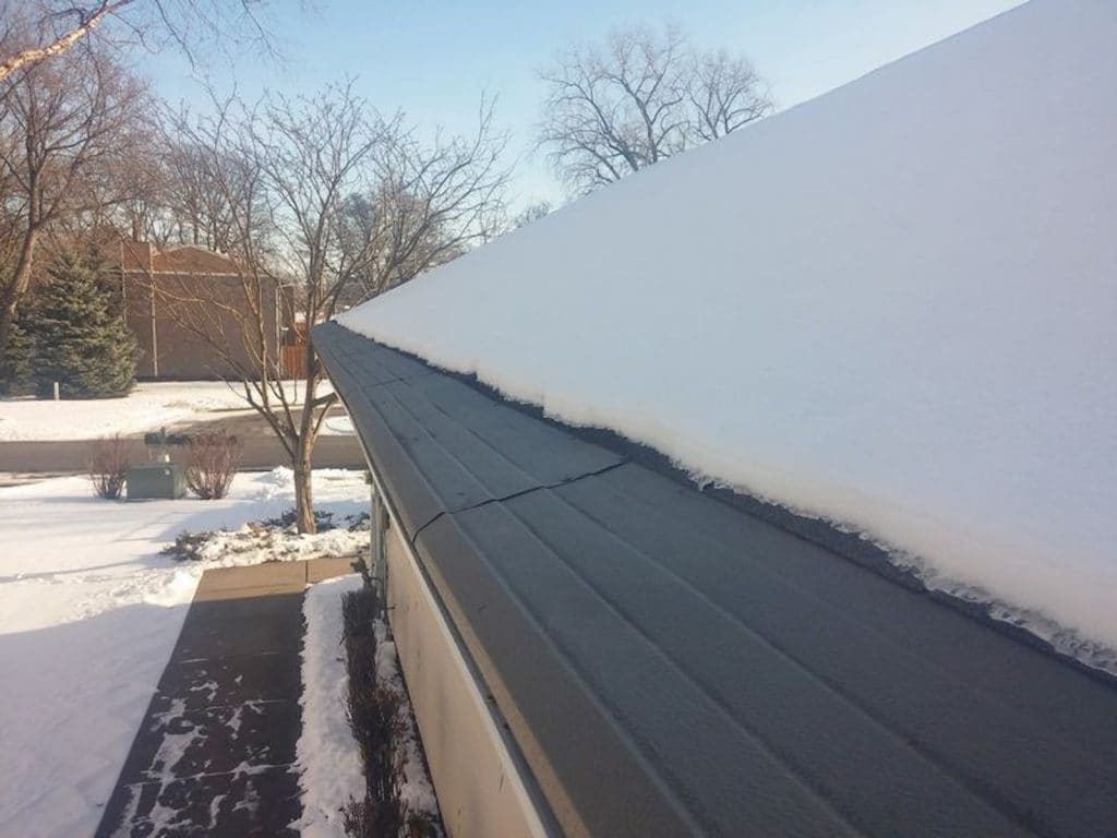 A metal roof edge with a thick, straight layer of snow resting along the top, set against a bright, sunny winter backdrop.
