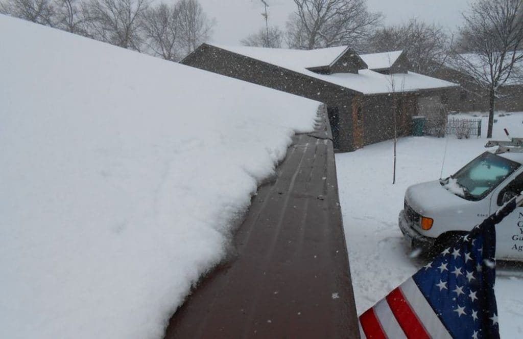 A thick layer of snow covers a roof edge, with a snowy yard, a van, and an American flag visible in the background.