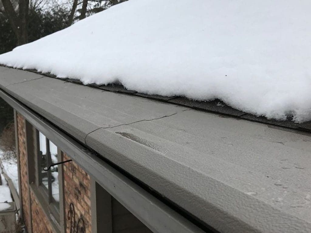 A thick layer of snow covers the shingled roof edge of a brick building with gray metal flashing and gutters.