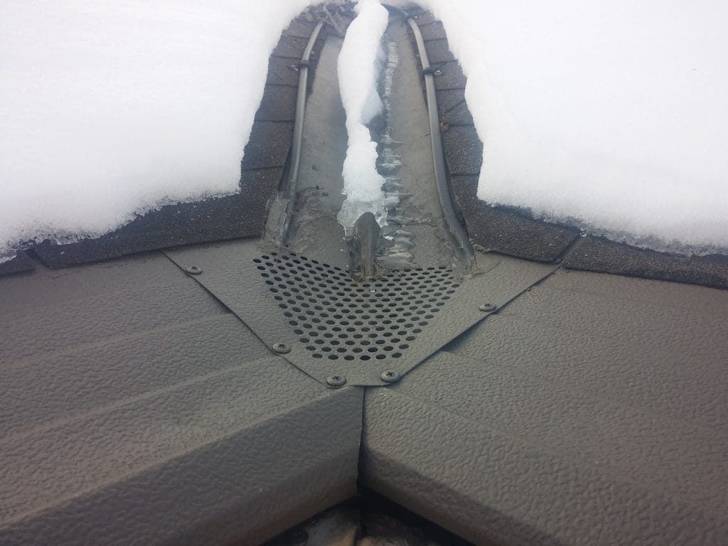 A snow-covered roof valley features a dark, perforated triangular drain guard secured with screws over the gutter joint.