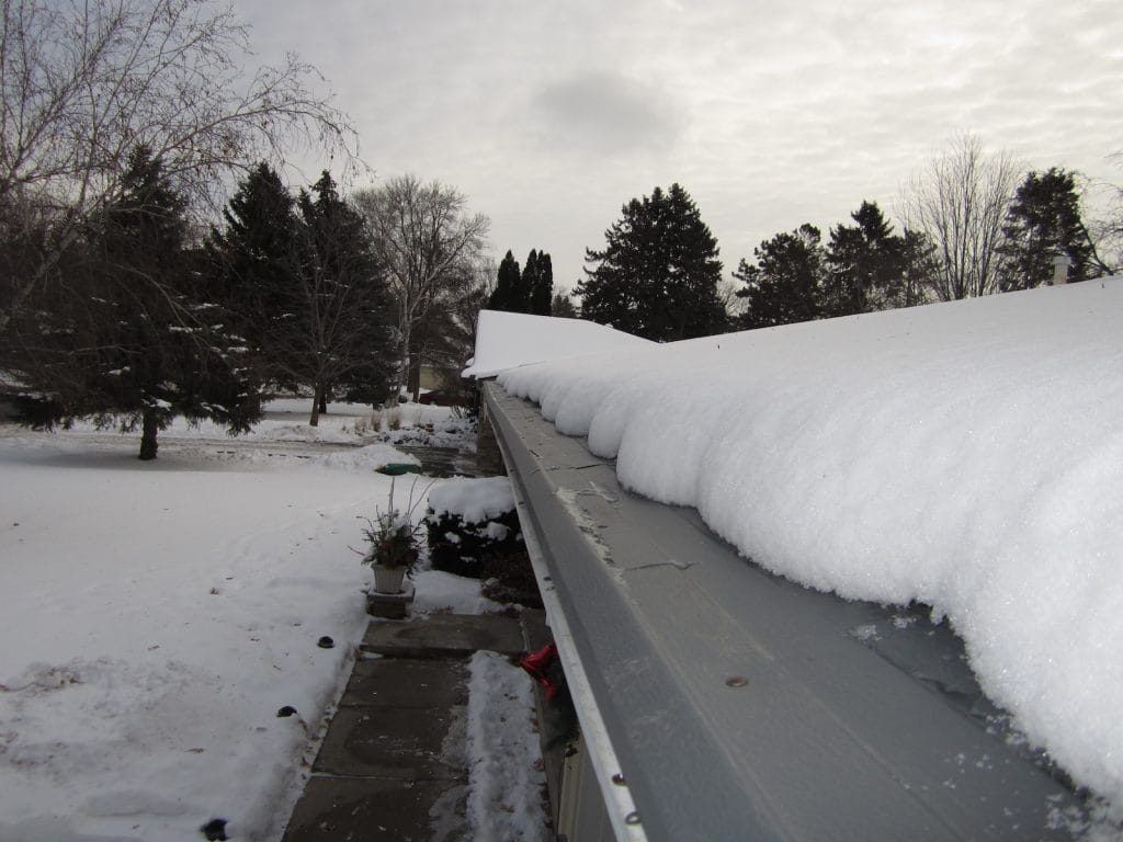 A thick layer of snow blankets the edge of a grey roof, with trees and a snow-covered yard visible in the background.