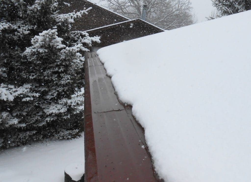 A close-up view of a metal roof gutter filled with snow, running along the edge of a snow-covered roof during a snowfall.