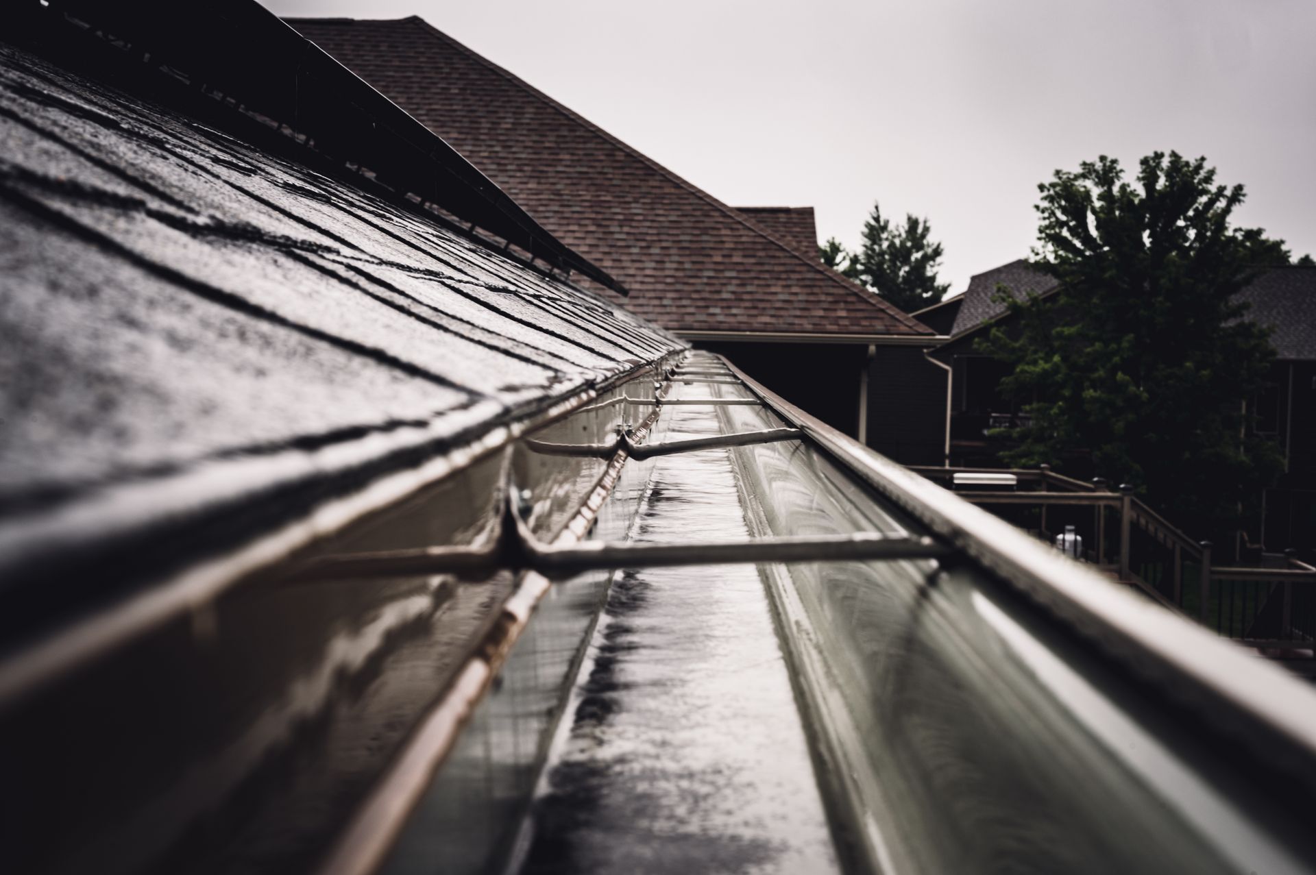 A close-up, angled view of a metal rain gutter attached to a shingled roof under a cloudy sky.
