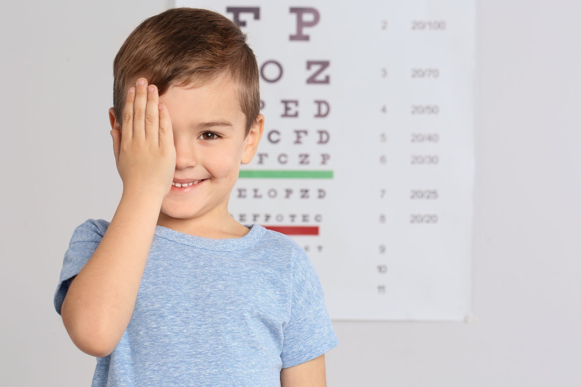 Photograph of a young boy covering one eye while standing in front of a Snellen eye chart used for