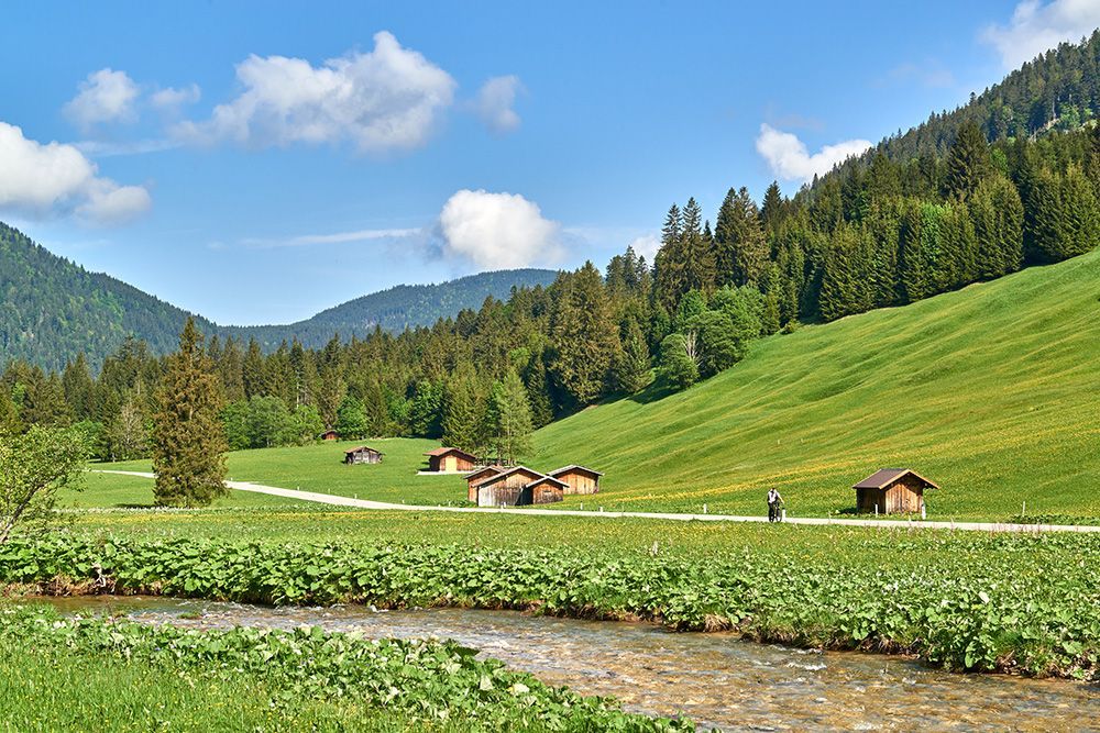 Idyllischer Bachlauf an grüner Almwiese mit alten Holzhütten und Radfahrer vor bewaldeten Bergen.