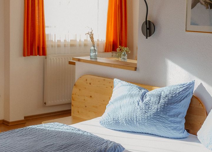 Helles Schlafzimmer mit Holzbett, hellblauer Bettwäsche und orangefarbenen Vorhängen am Fenster.
