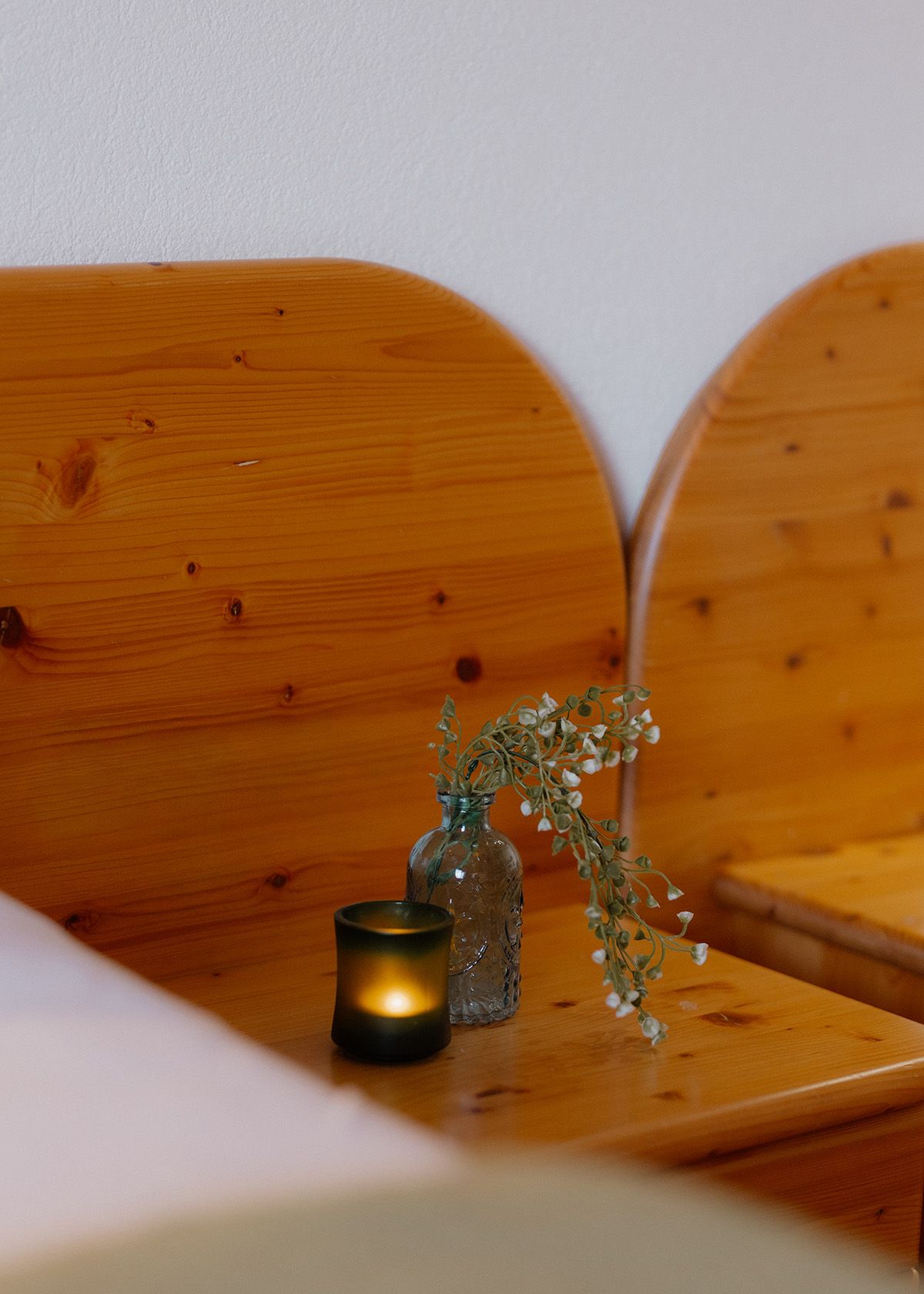 Holz-Nachttisch mit brennender Kerze und kleiner Blumenvase vor dem hölzernen Bettkopfteil.