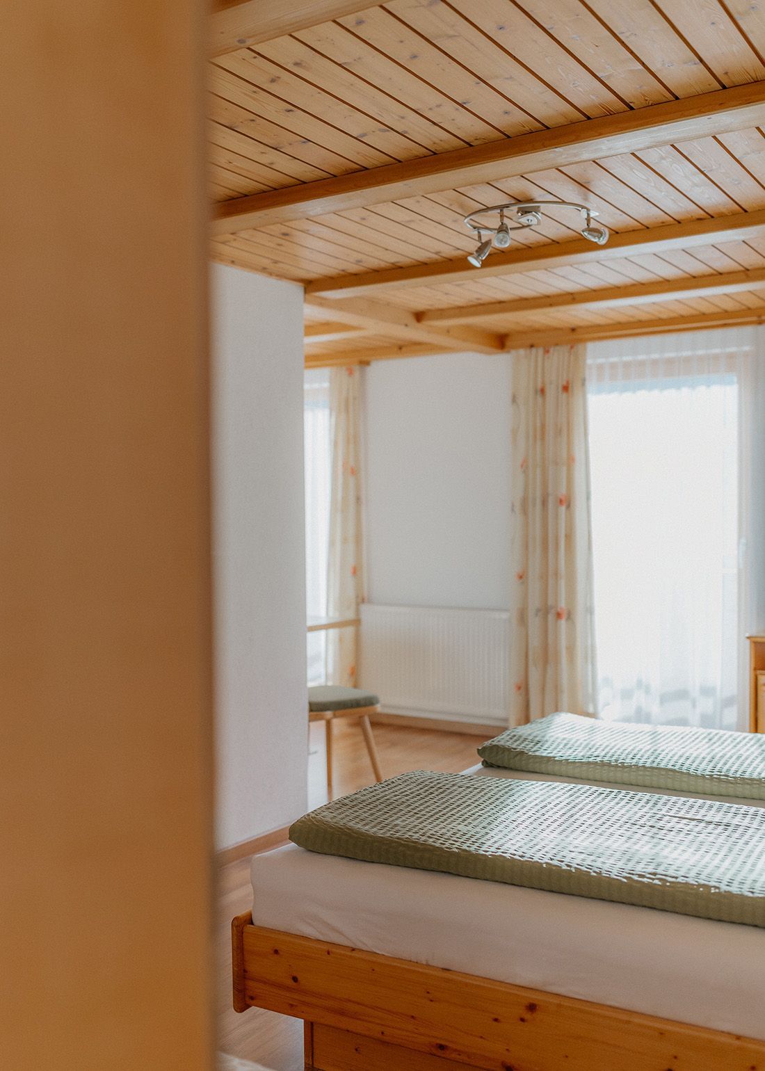 Helles Schlafzimmer einer Ferienwohnung mit rustikaler Holzdecke und gemütlichem Holzbett mit grüner Decke.