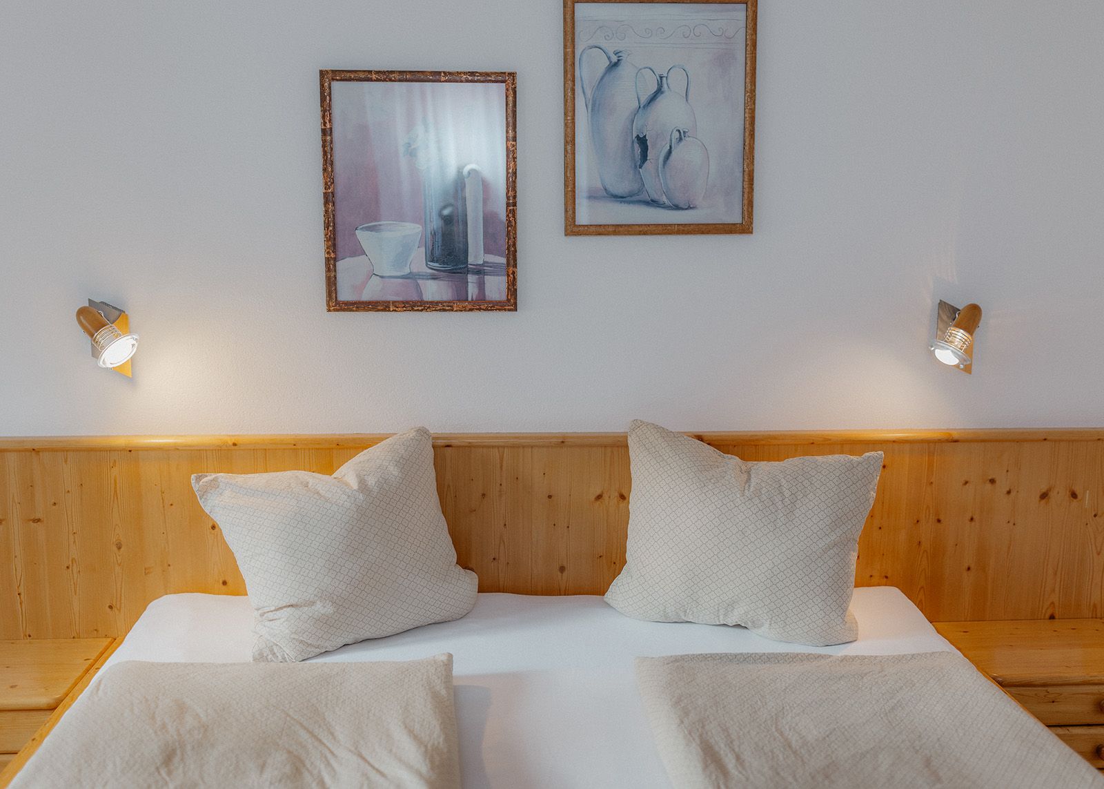 Holz-Doppelbett mit hellen Kissen, Leselampen und Wandbildern im gemütlichen Schlafzimmer einer Ferienwohnung.