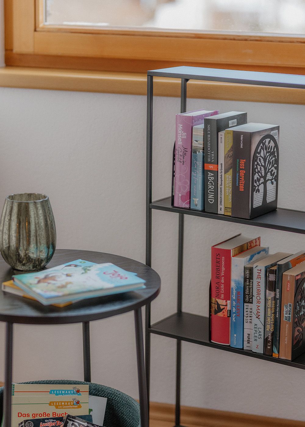 Schwarzes Metall-Bücherregal mit Romanen neben einem runden Beistelltisch in einer gemütlichen Leseecke.