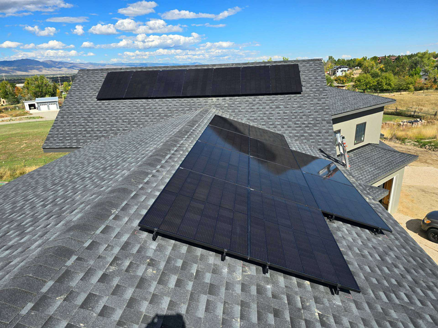 Solar panels installed on a gray shingled roof under a blue sky, with a rural landscape in the background.