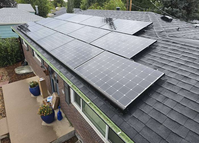 Solar panels installed on a dark-shingled roof of a brick house. Green trim, blue pots, and a cloudy sky visible.