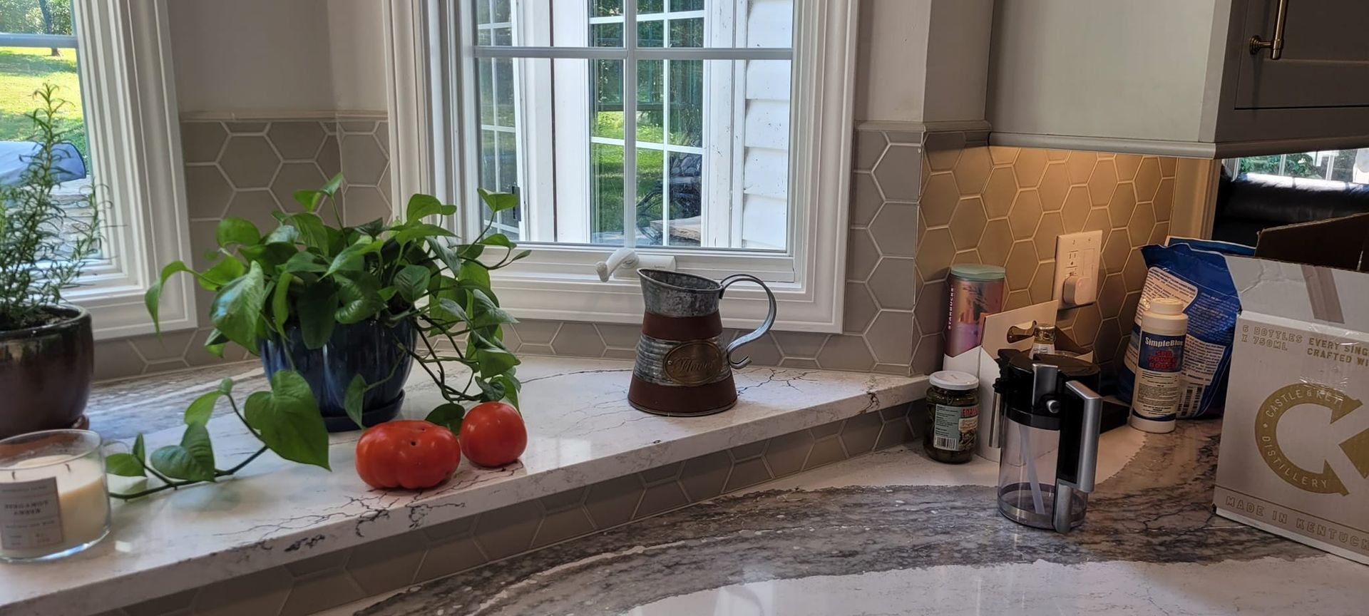 A kitchen window sill with plants, tomatoes, and a pitcher.