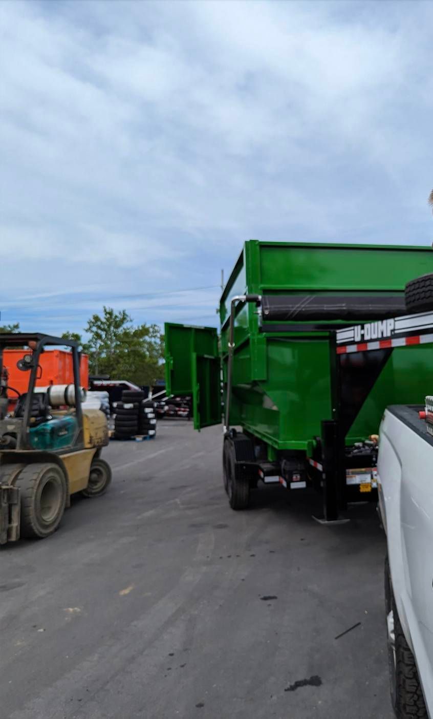 Green dump truck with open sides parked on asphalt, a forklift on the left. Cloudy sky.