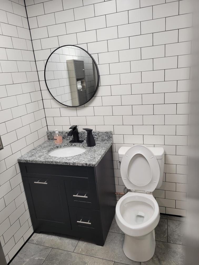 Bathroom with white brick tile walls, dark gray vanity, round mirror, and open toilet.
