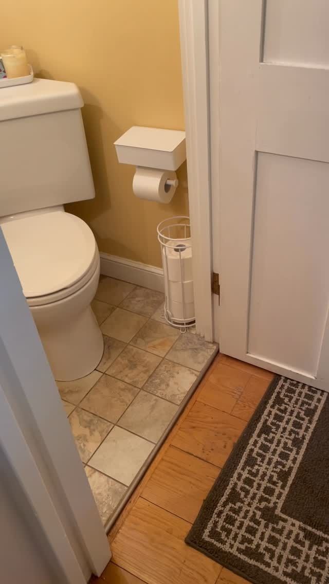 Small bathroom with toilet, toilet paper holder, trash can, and open door by a rug on tiled and wood floor.