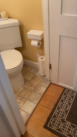 Small bathroom with toilet, toilet paper holder, trash can, and open door by a rug on tiled and wood floor.