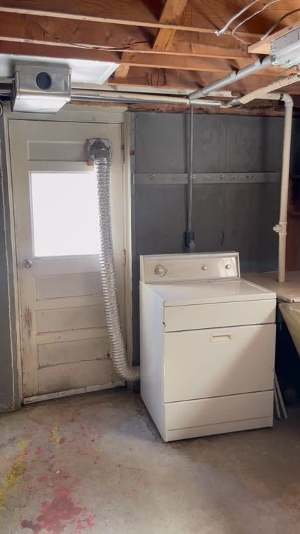 Laundry area with a white dryer beside a door, exposed ceiling beams, and unfinished walls.