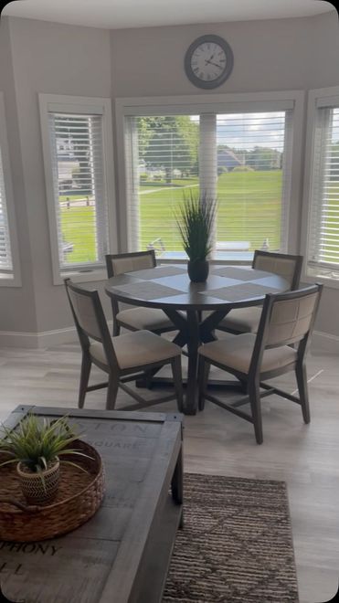 Dining nook with round table, four chairs, tall plant, bay windows, and green lawn outside.