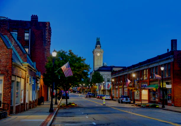 Quiet small-town street at dusk with lit storefronts, an American flag, and a clock tower in the distance