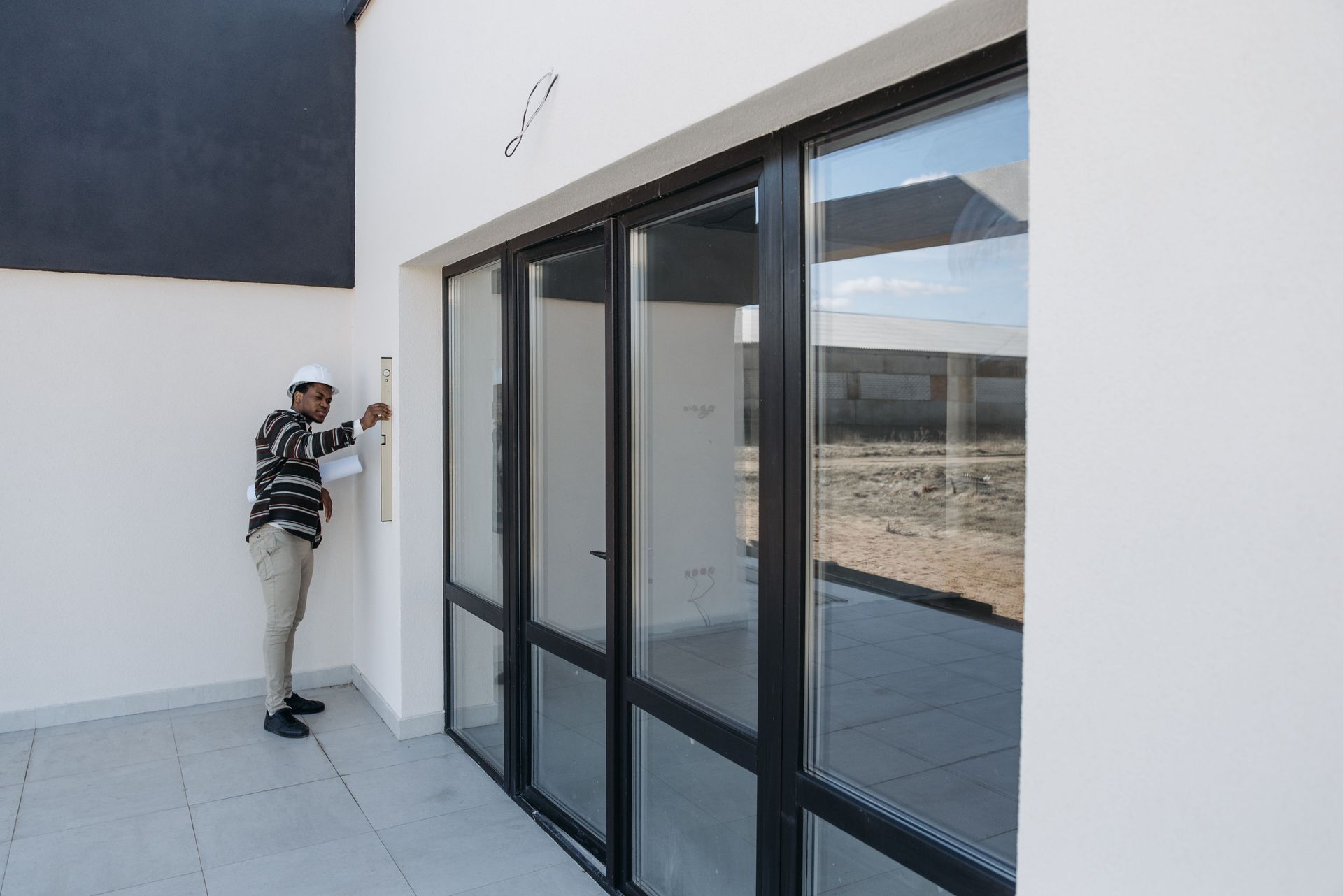 Person standing beside a white building near large glass doors on a sunny patio.