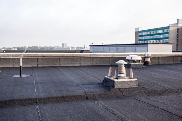 A flat, grey gravel-surfaced roof with ventilation pipes and a concrete perimeter wall, against a distant cityscape.