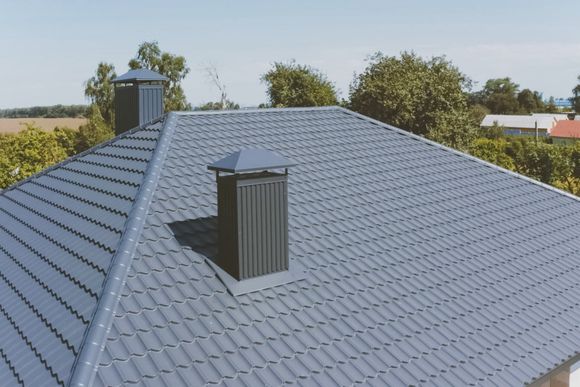 A gray, tiled hip roof featuring two dark, rectangular chimneys with matching caps against a backdrop of trees and sky.