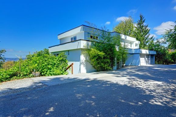 A white, modern, two-story house with a flat roof, surrounded by green shrubs, situated under a bright blue sky.