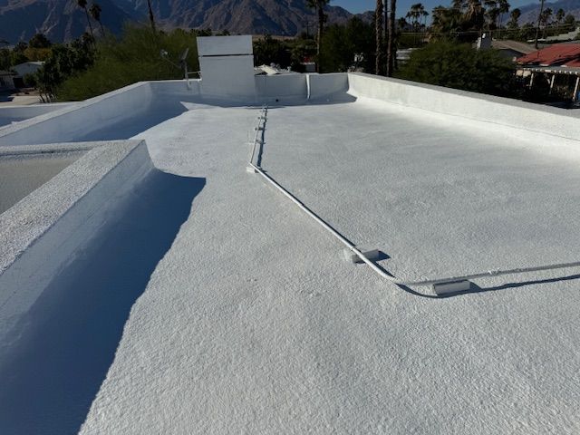 A wide-angle, high-angle shot of a flat roof coated in white, textured sealant with conduit running across the surface.