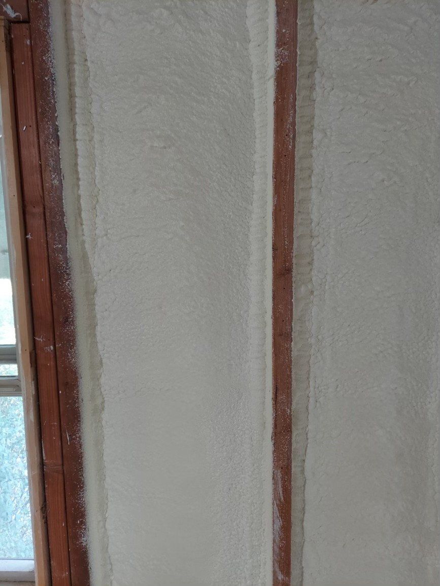 Light-colored spray foam insulation applied between vertical wooden wall studs.