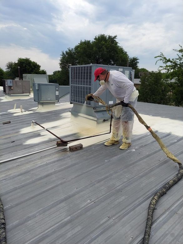 A worker in protective gear applies spray foam insulation to a metal roof near an HVAC unit on a cloudy day.