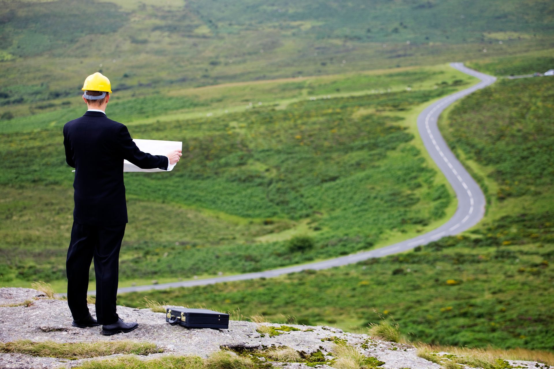 A man in a hard hat is standing on top of a hill looking at a road.