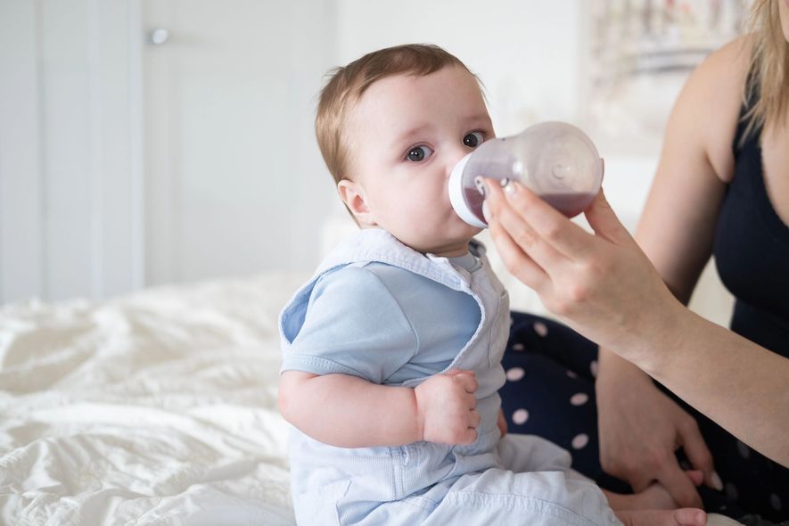 Un bambino beve dal biberon seduto su un letto accanto al braccio di un adulto.